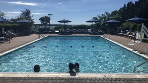 Piscina con sillones de descanso y sombrillas, rodeada de árboles y un cielo despejado.
