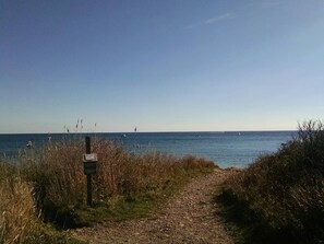 Un sentier de gravier mène à une plage d’où l’on a une vue dégagée sur la mer et sur les bateaux qui se trouvent au loin.