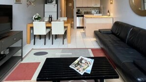 Una sala de estar moderna con un sofá negro, un comedor con una mesa y sillas de madera, una cocina con gabinetes blancos y un pequeño balcón con barandilla blanca.