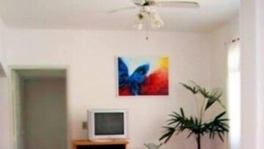 Una habitación con ventilador de techo, televisión sobre un mueble de madera, una planta y una silla roja.