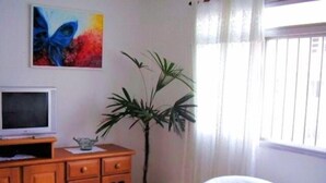 Una sala de estar con un televisor sobre un mueble de madera, una planta en maceta, una silla blanca y un cuadro de colores en la pared.