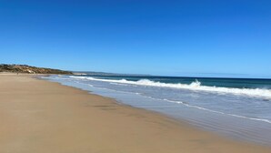 A wide sandy beach with gentle waves, a clear blue sky, and a distant hill with buildings.