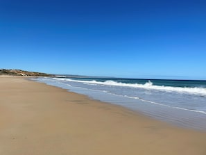 A wide sandy beach with gentle waves, a clear blue sky, and a distant hill with buildings.