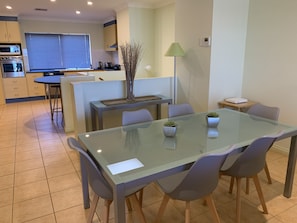 A modern kitchen with built-in appliances, a dining area with a glass table and chairs, and a small side table with a lamp.