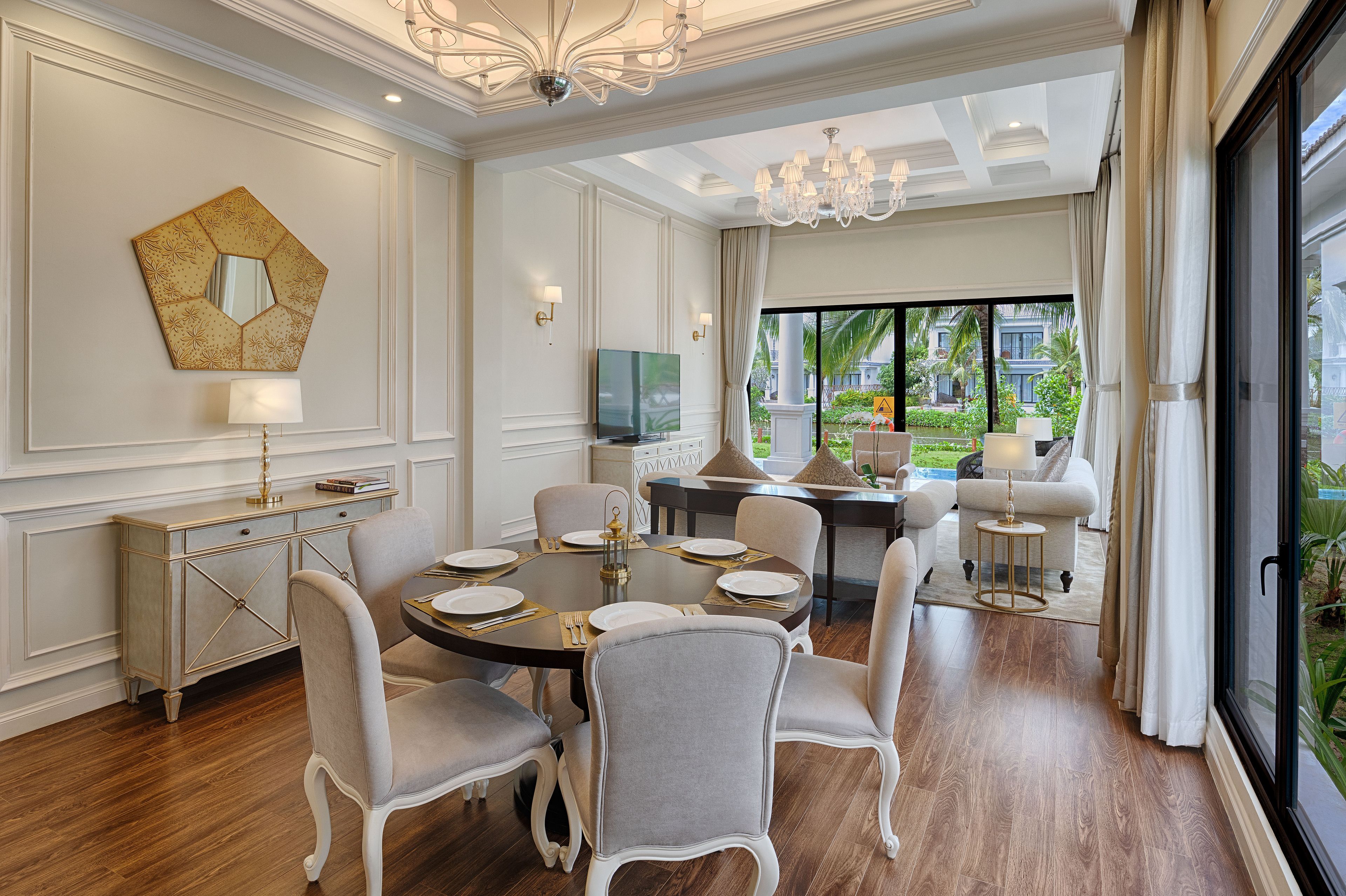 A dining area with a round table set for six, surrounded by six chairs. There is a television on the wall and a large window providing a view of the outdoors.