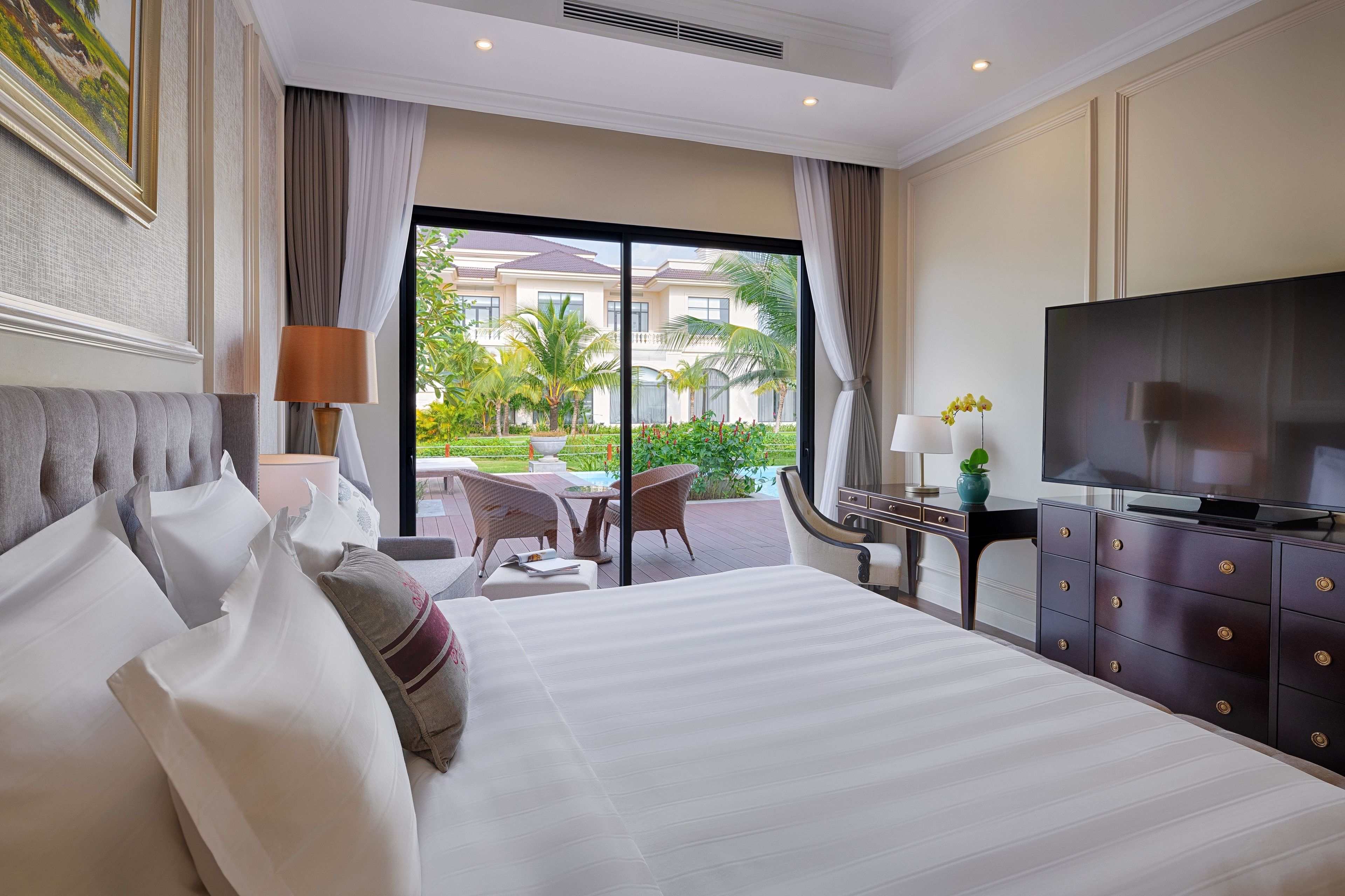A hotel room with a large bed, a television, a desk with a lamp, and a view of a landscaped area outside.