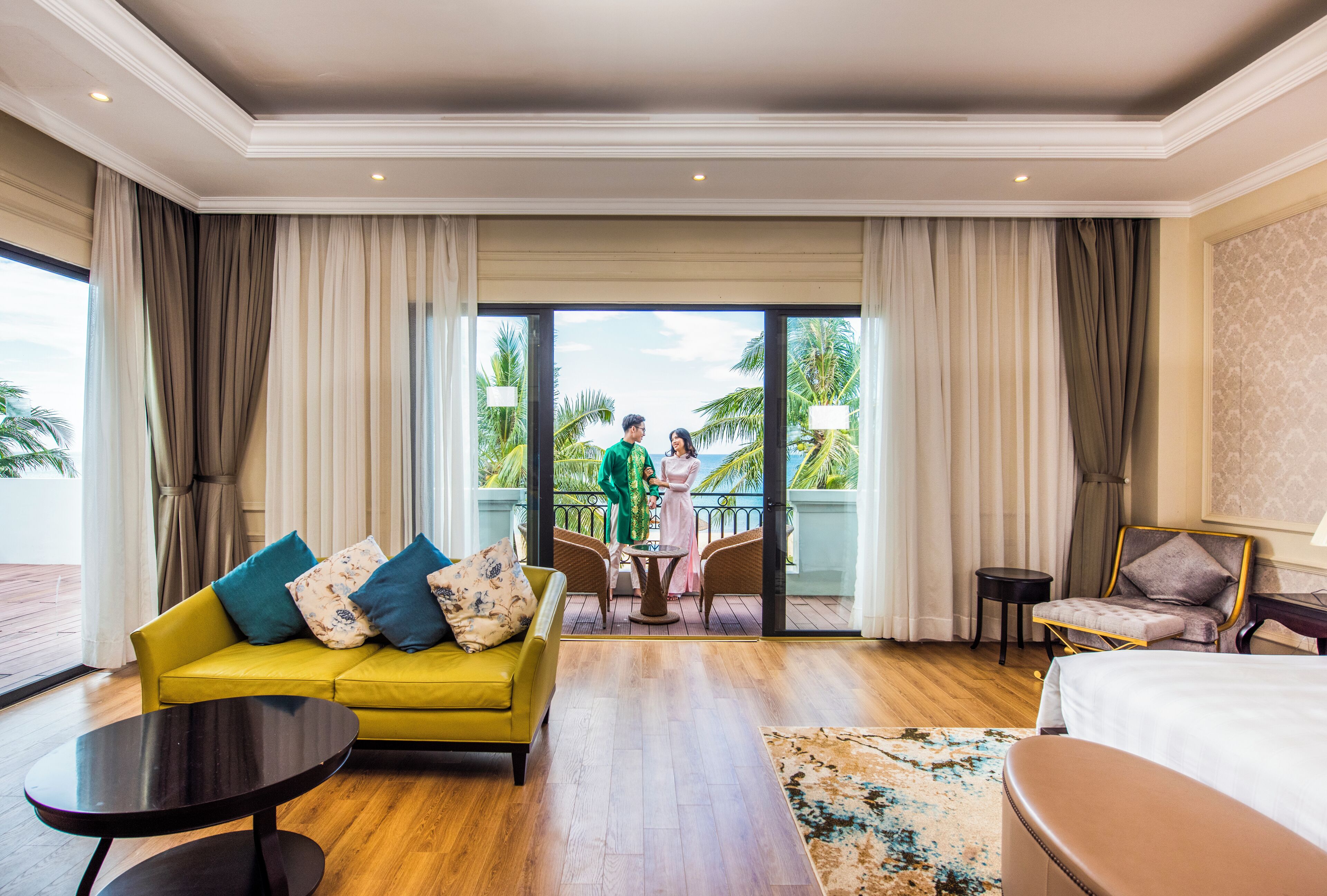 Habitación de hotel con sofá amarillo, una mesa redonda, cama y balcón con vista a palmeras.