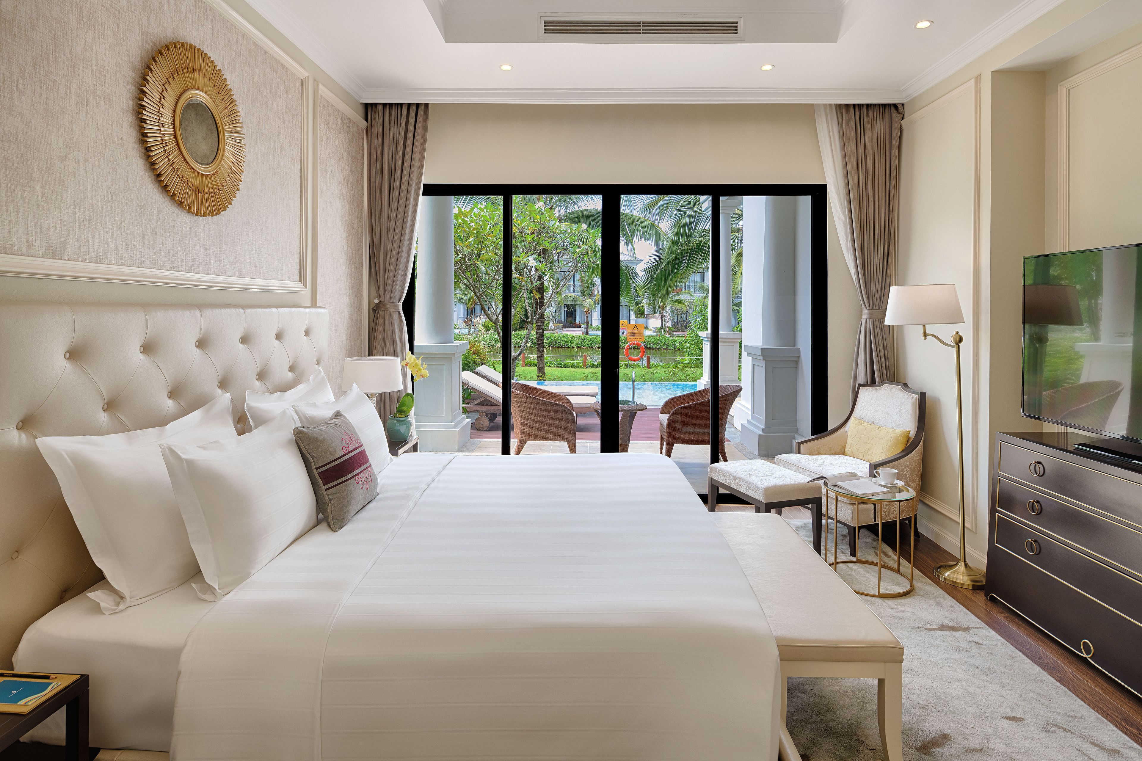 A large bed with a tufted headboard, a television, a desk, and a view of a pool and greenery outside.