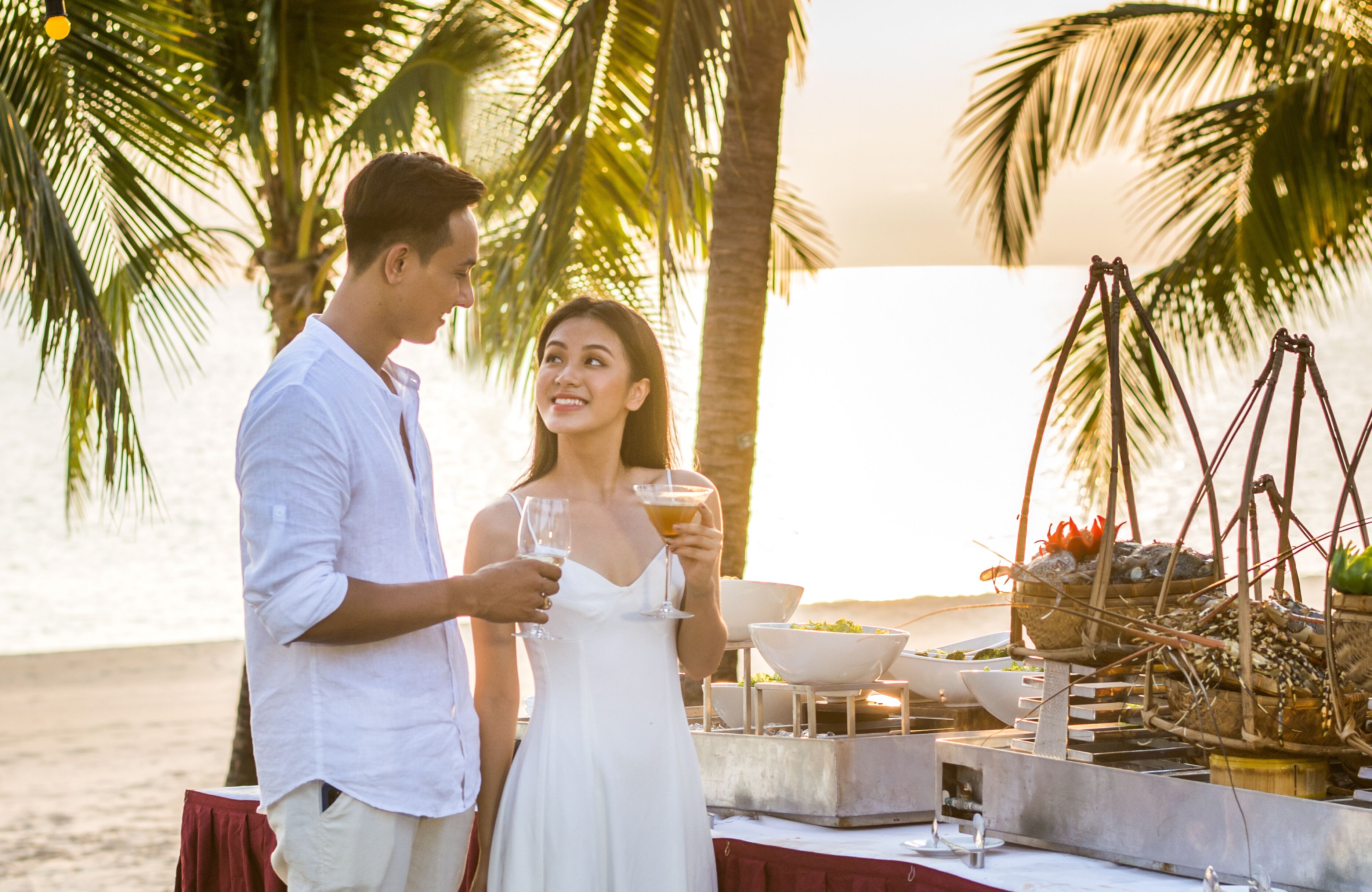 A couple standing by a table with a tropical setting, including palm trees and a sunset in the background.