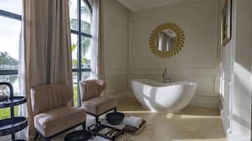 Un baño con una bañera independiente, dos sillones, un espejo grande y una ventana con cortinas.