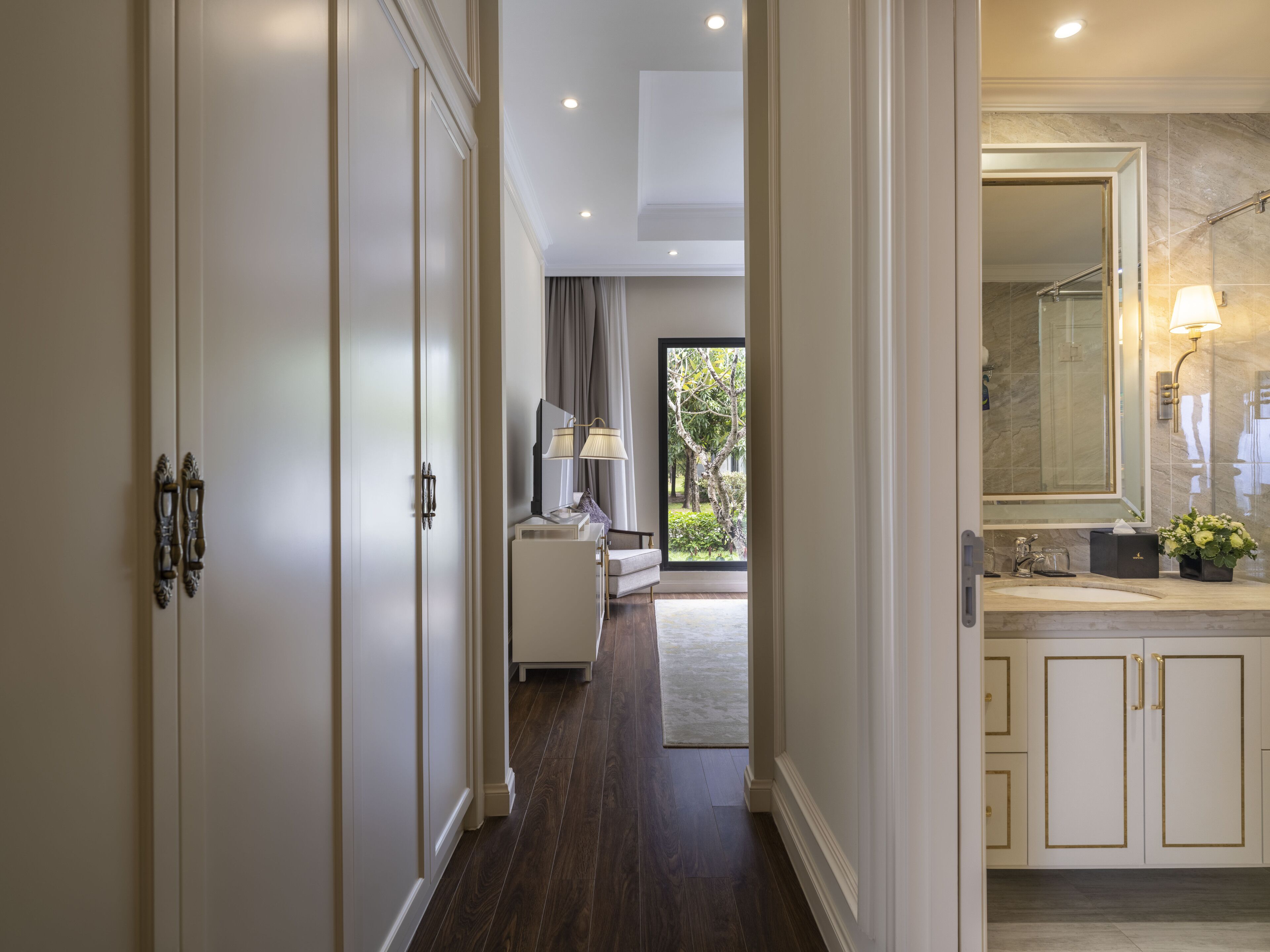 Un couloir au plancher de bois mène à une chambre à coucher qui contient un lit et une fenêtre. On y trouve également une salle de bain avec un lavabo et un miroir.