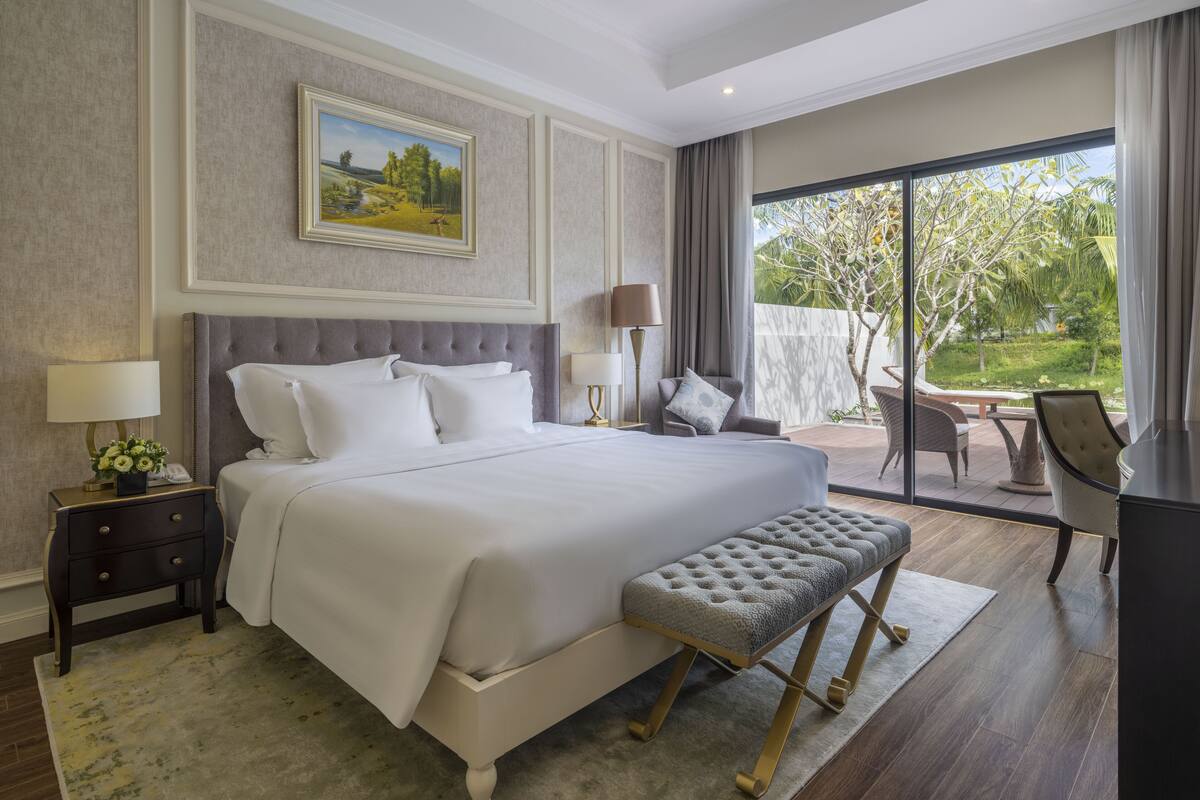 Ruime slaapkamer met een groot bed, een bankje, een bureau en uitzicht op een tuin door een schuifdeur.