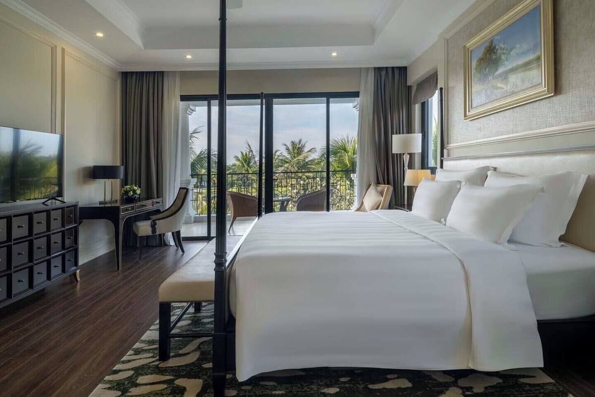 Hotelkamer met een groot bed, een bureau met televisie, een balkon met uitzicht op palmbomen en een plafondventilator.