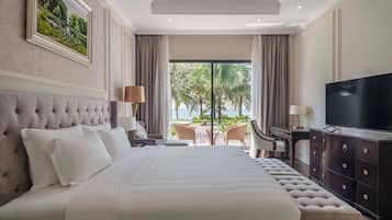 A large bed with a tufted headboard, a flat-screen TV, a desk with a lamp, and a view of the ocean through large windows.