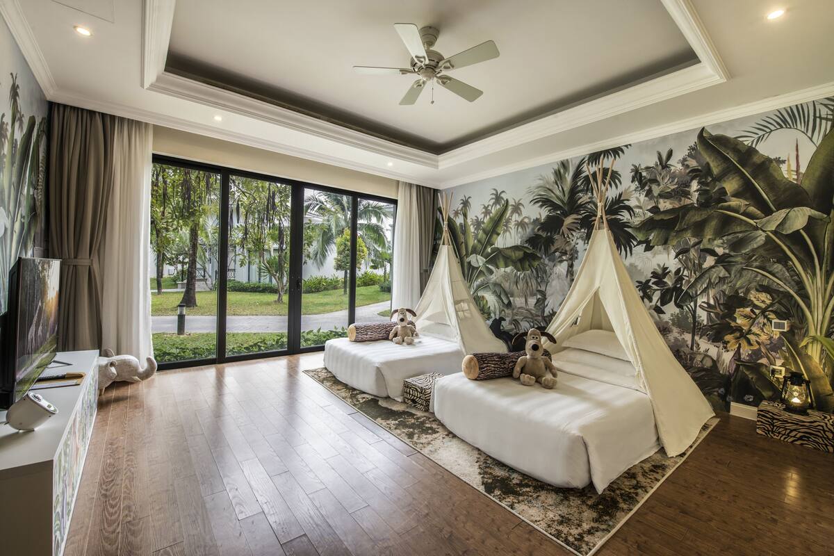 Een ruime kamer met een groot raam, een plafondventilator, een muurschildering van tropisch groen en een bedstructuur in tentstijl.
