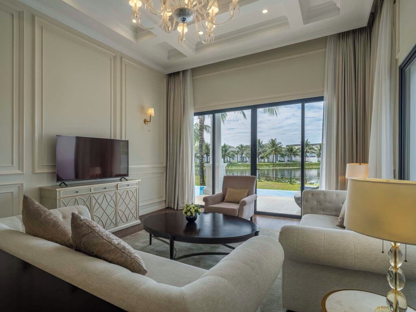 A spacious living room with a large flat-screen TV, a sofa, two armchairs, a coffee table, and a view of a pool and palm trees through large windows.