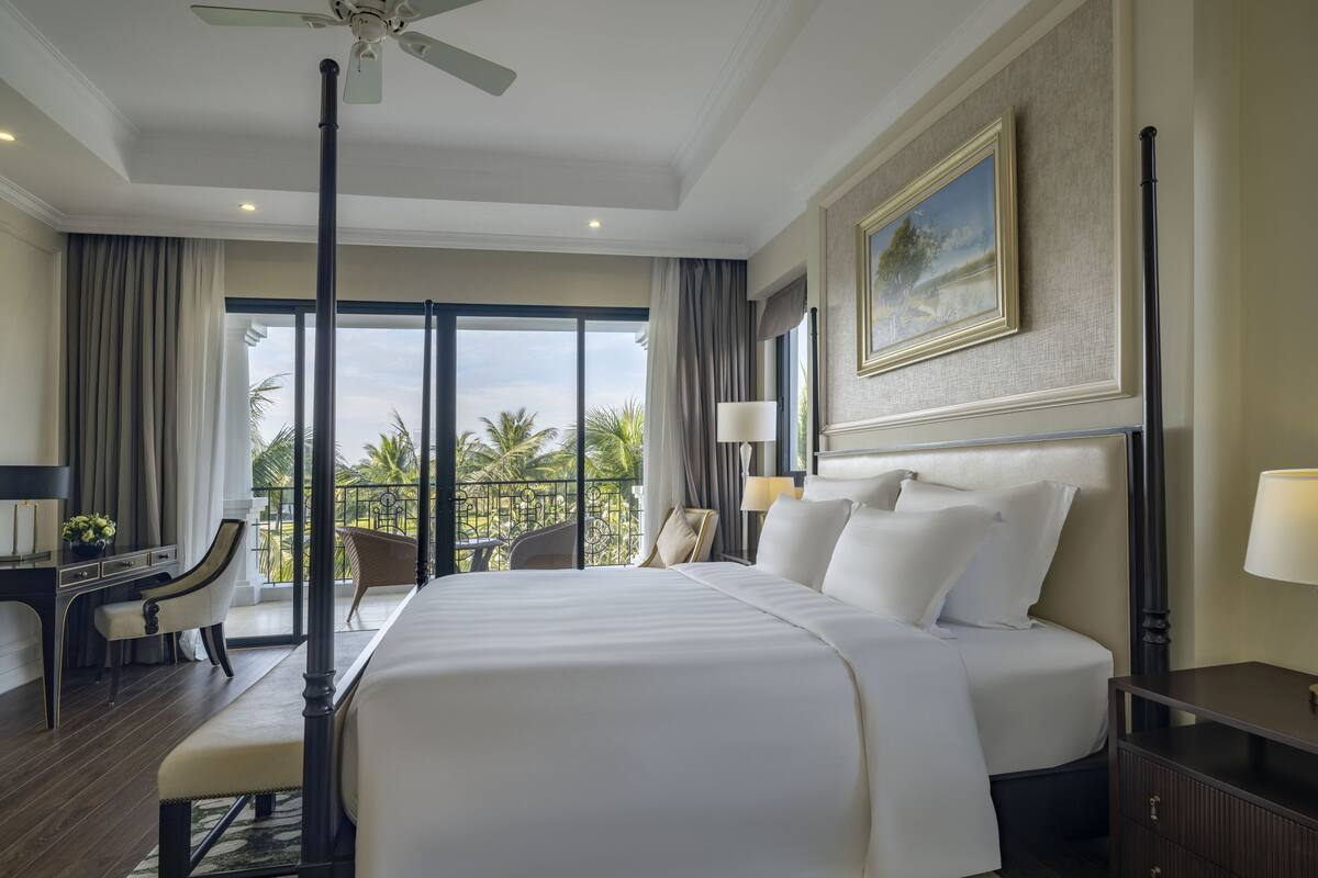 Een hotelkamer met een groot bed, een bureau met stoel, een balkon met uitzicht op palmbomen en een plafondventilator.