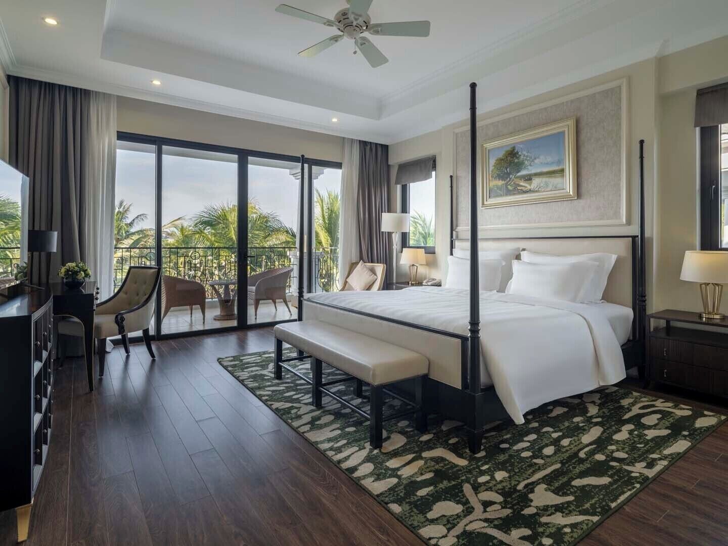 Habitación de hotel con cama con dosel, escritorio con silla, balcón con vista a palmeras y un cuadro en la pared.