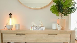 Une commode en bois avec un miroir au-dessus, une lampe, une plante en pot et une pile de livres.