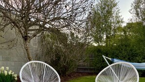 Un foyer circulaire avec des flammes, entouré de blocs de pierre, et deux chaises blanches en fil de fer, le tout dans un jardin.