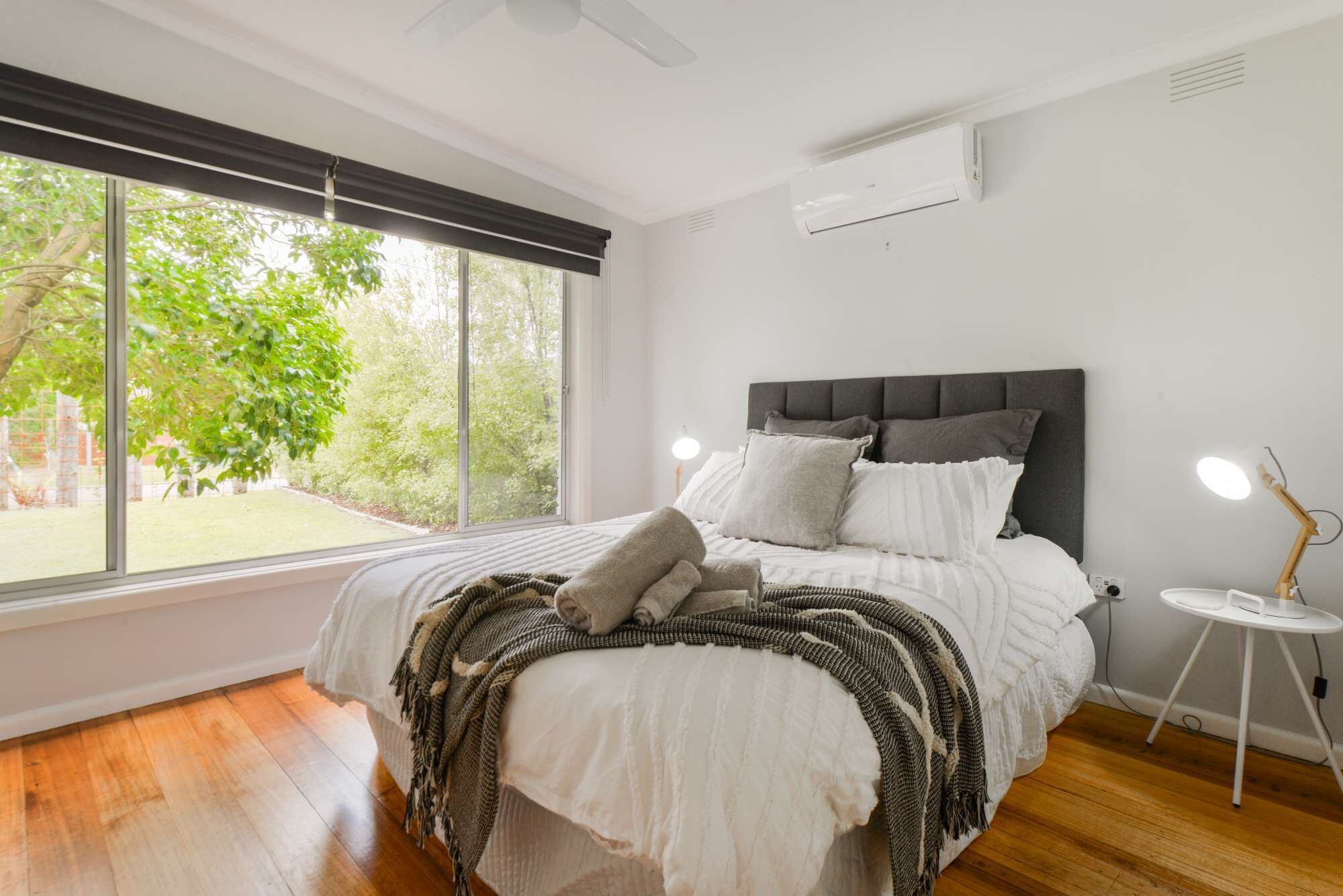 A bedroom with a large window, a bed with white bedding, a grey headboard, and a bedside table with a lamp.