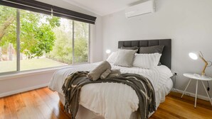 Une chambre avec une grande fenêtre, un lit avec une literie blanche, une tête de lit grise et une table de chevet avec une lampe.