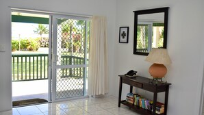 A room with a sliding glass door leading to a balcony, a small table with a lamp and books, and a framed picture on the wall. - Paere House, Snorkeling Heaven (Tikioki Beach)