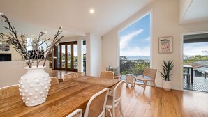 A modern dining area with a wooden table and white chairs, a white vase with branches, and a view of the sea through large windows. - Pet Friendly - Bay Views From Drury Lane with Solar Heated Pool and Gas Heated Spa (Dromana)