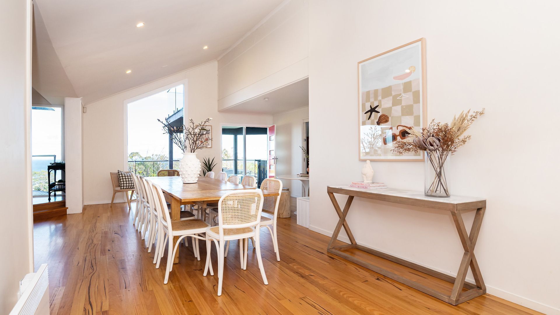 A spacious dining area with a wooden table and white chairs, a console table with a vase, and a large window providing natural light.
