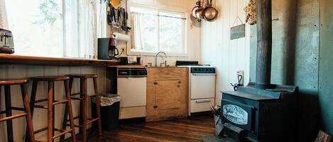 A rustic kitchen with wooden stools, a stove, and a window overlooking greenery.