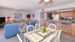 A living room with a sofa, chairs, a dining table with a vase of flowers, and a kitchen area with cabinets and appliances. - Direct Gulf Front Beach Paradise! Any Closer To The Water & You Would Be In It.. (Indian Shores)