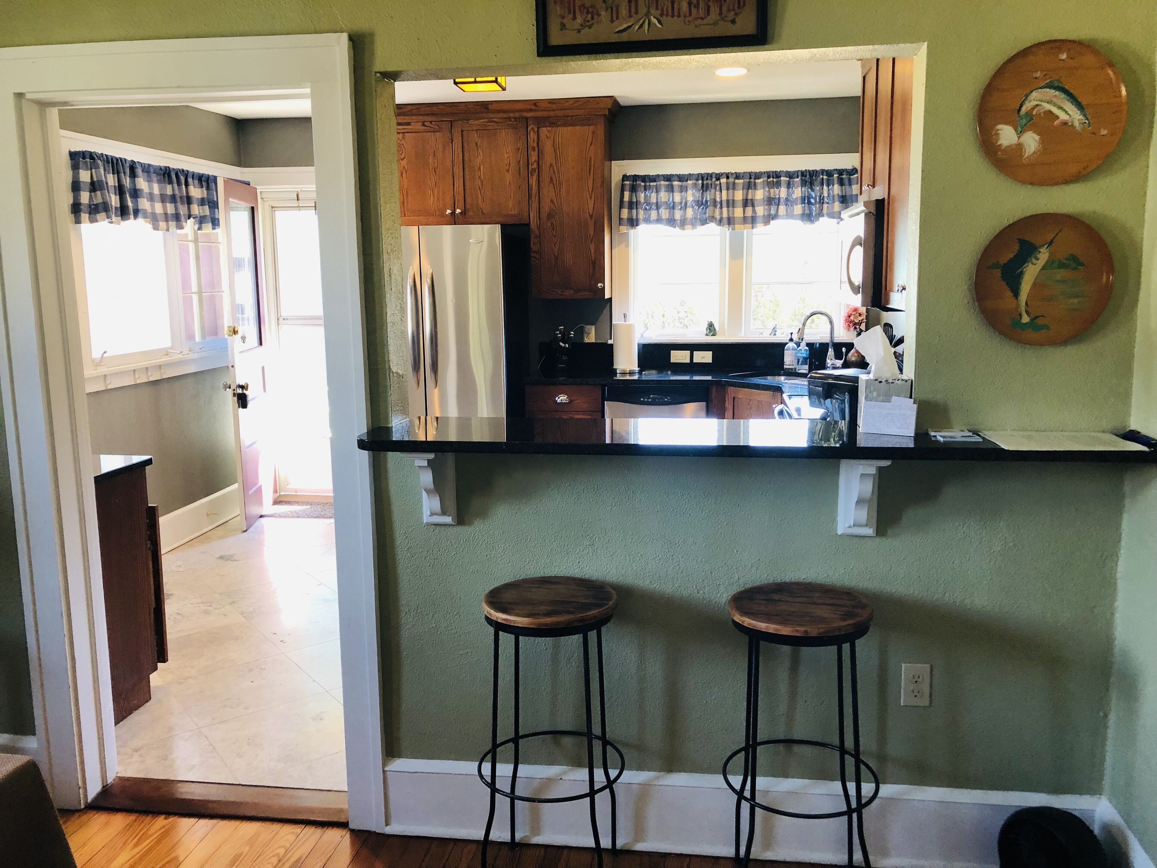 A kitchen with wooden cabinets, a stainless steel refrigerator, a black countertop, and two stools.
