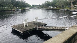 Un muelle de madera con sillas de plástico blanco que se extiende sobre aguas tranquilas, rodeado de árboles y algunos barcos.
