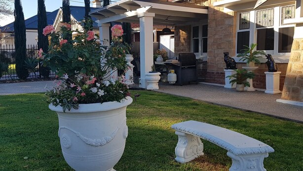 Un jardín con una jardinera grande y blanca llena de flores rosadas, un banco blanco y una zona de patio con una parrilla y mobiliario exterior.