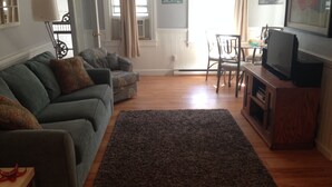 Ein Wohnzimmer mit grauem Sofa, einem dunklen Teppich, einem Holz-TV-Möbel und einem Essbereich mit Tisch und Stühlen.