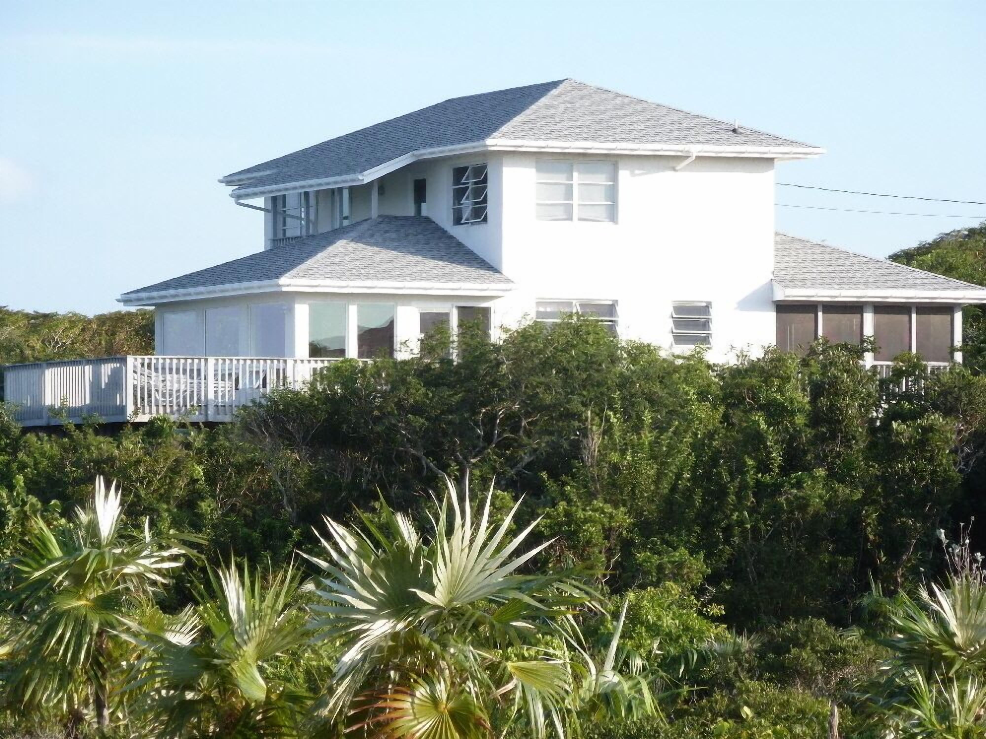 Une maison à deux étages, à l’extérieur blanc et au toit gris, entourée d’une végétation luxuriante et de palmiers.