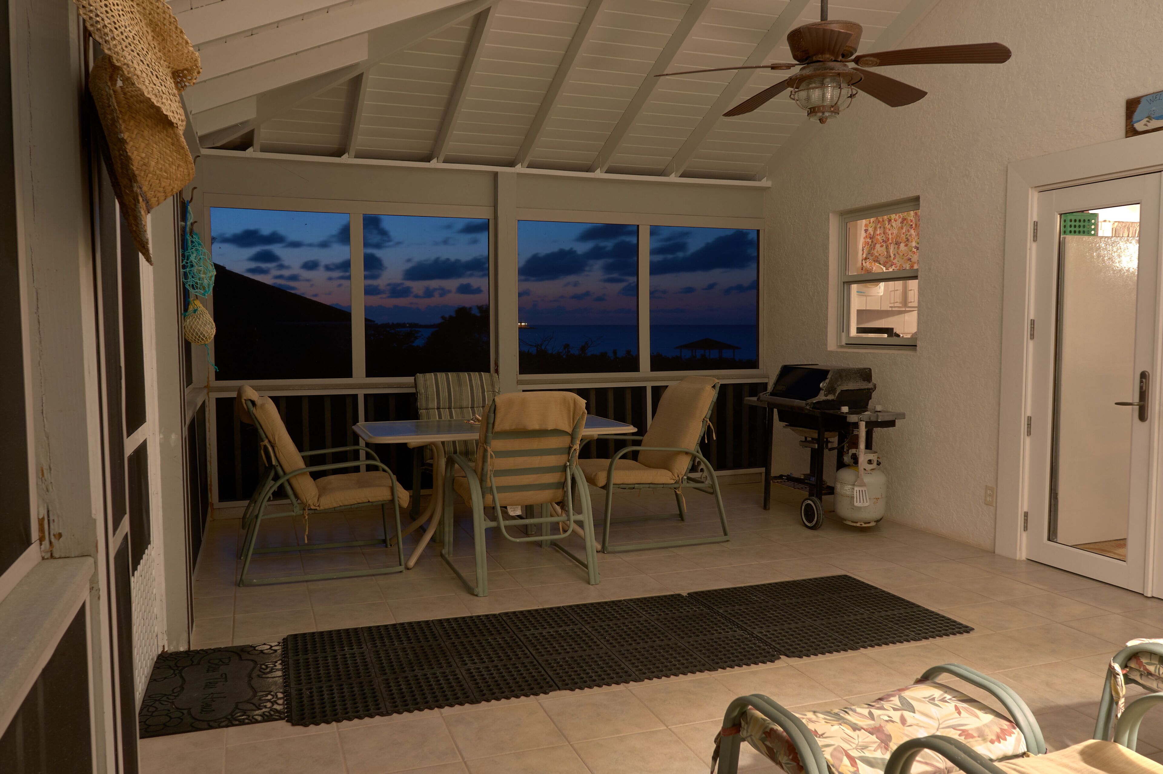 Um alpendre coberto com mesa e cadeiras, um ventilador de teto e vista para o oceano ao entardecer.