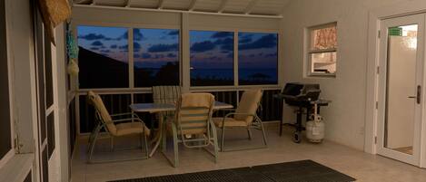 Um alpendre coberto com mesa e cadeiras, um ventilador de teto e vista para o oceano ao entardecer.