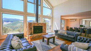 A spacious living room with a stone fireplace, large windows offering a mountain view, a dark leather sofa, and a dining area with a kitchen in the background.