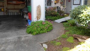 Un garage con pavimento in cemento, porta in legno e un piccolo giardino con fiori rosa e piante verdi.