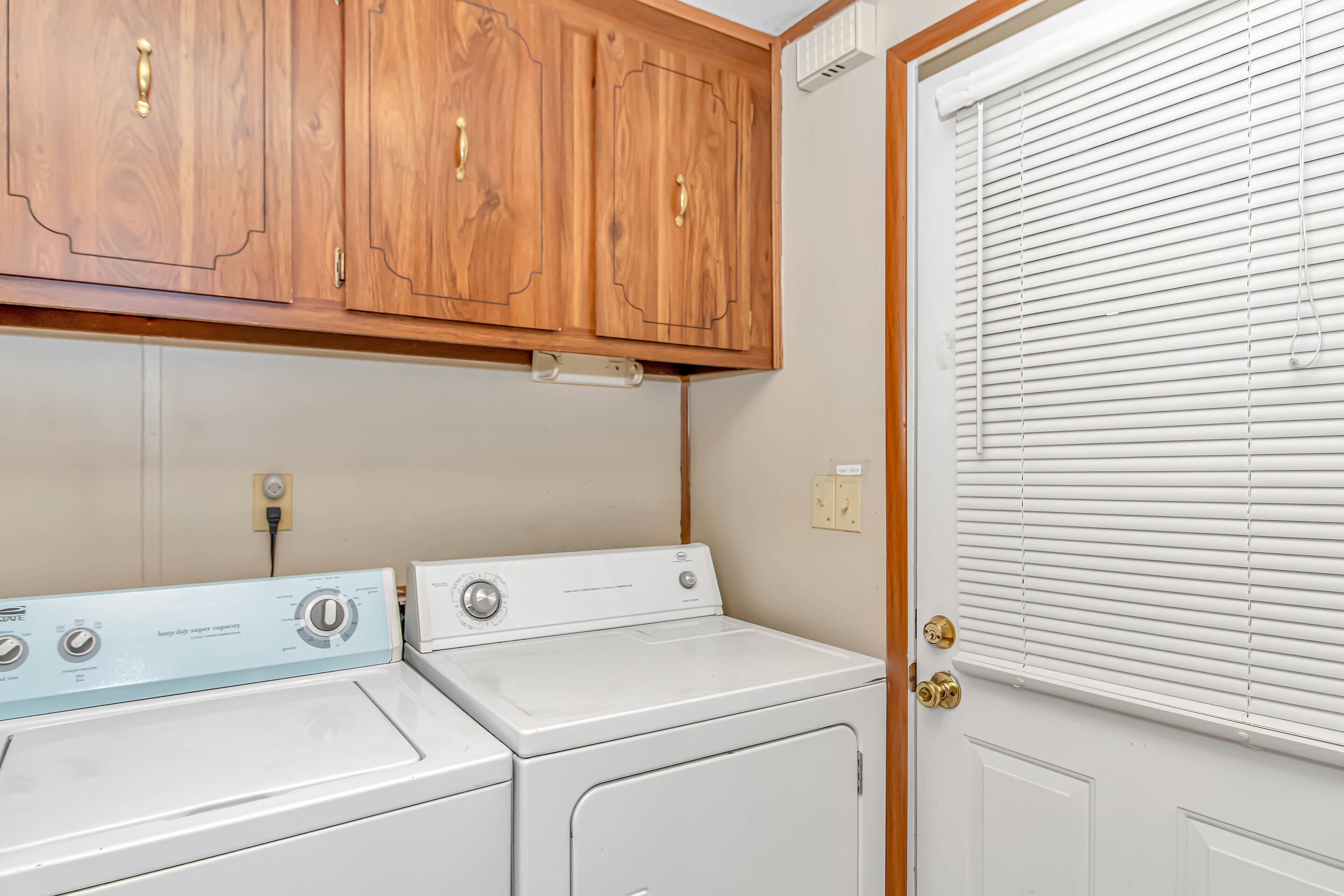 A laundry room with two white washers and dryers, wooden cabinets above, and a door with blinds.