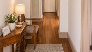 Un pasillo con piso de madera, un arco de entrada, cuadros en las paredes, una mesa con una lámpara y un cartel de bienvenida, y una planta en maceta.