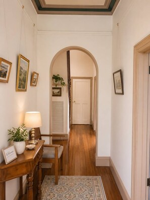 Un pasillo con suelo de madera, un arco de entrada, cuadros en las paredes, una mesa con lámpara y un cartel de bienvenida, y una planta en maceta.