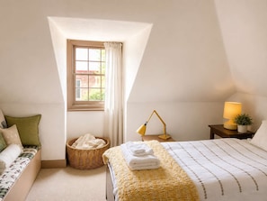 Ein Schlafzimmer mit einem Bett, einem Nachttisch mit Lampe, einem Fenster mit Vorhängen, einem Korbwäschekorb und einer kleinen Topfpflanze.