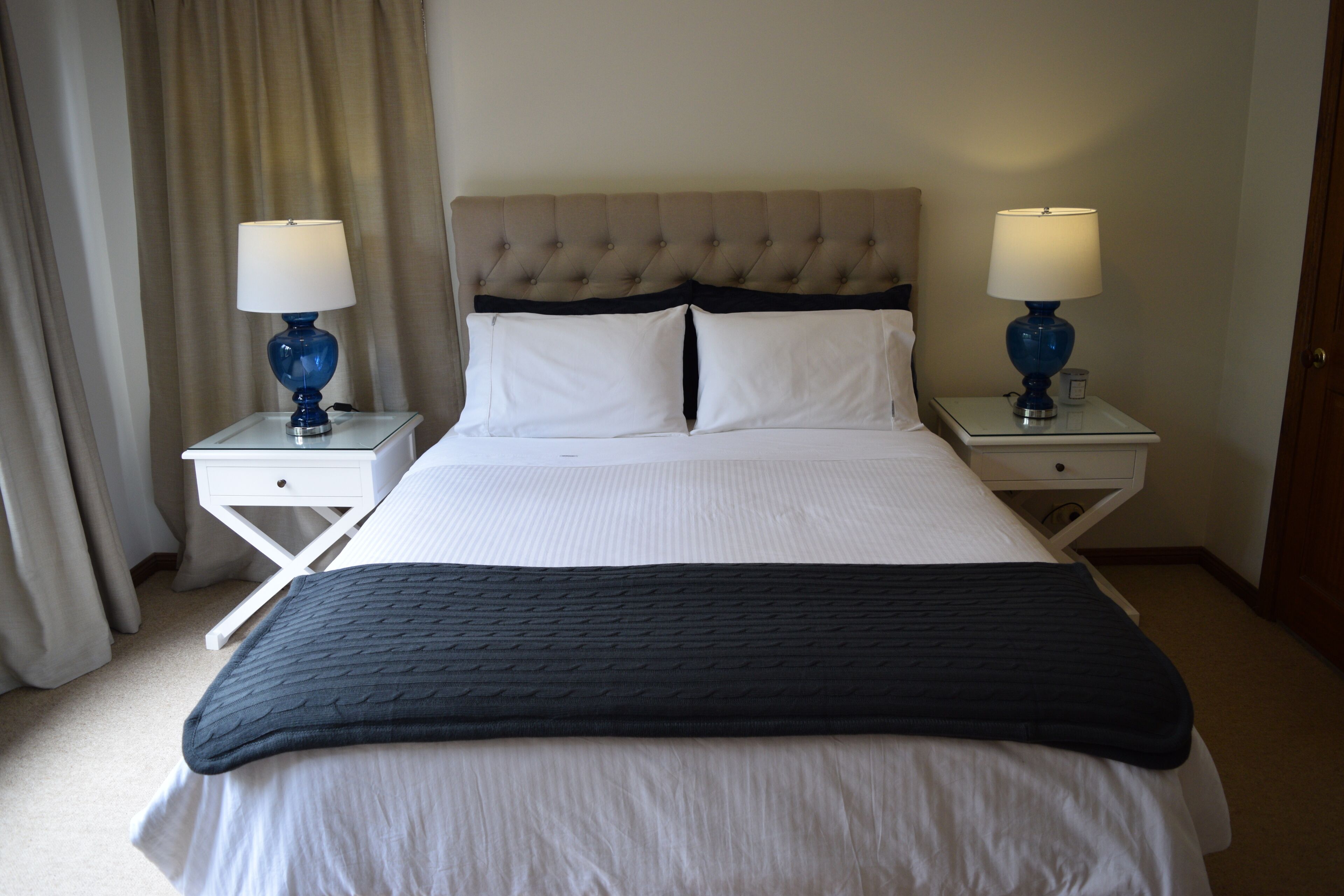 Un lit bien fait, recouvert d’une couvre-lit blanche et bleu foncé, avec deux lampes bleues assorties posées sur des tables de chevet blanches, et une tête de lit capitonnée.