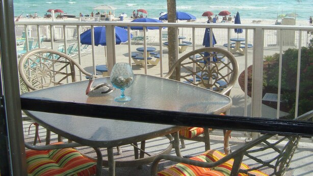 A balcony with a table and chairs, overlooking a beach with umbrellas and palm trees. - Oceanfront 1BR/2BR Condo on 2nd Floor, 1,000 SQ FT (Panama City Beach)