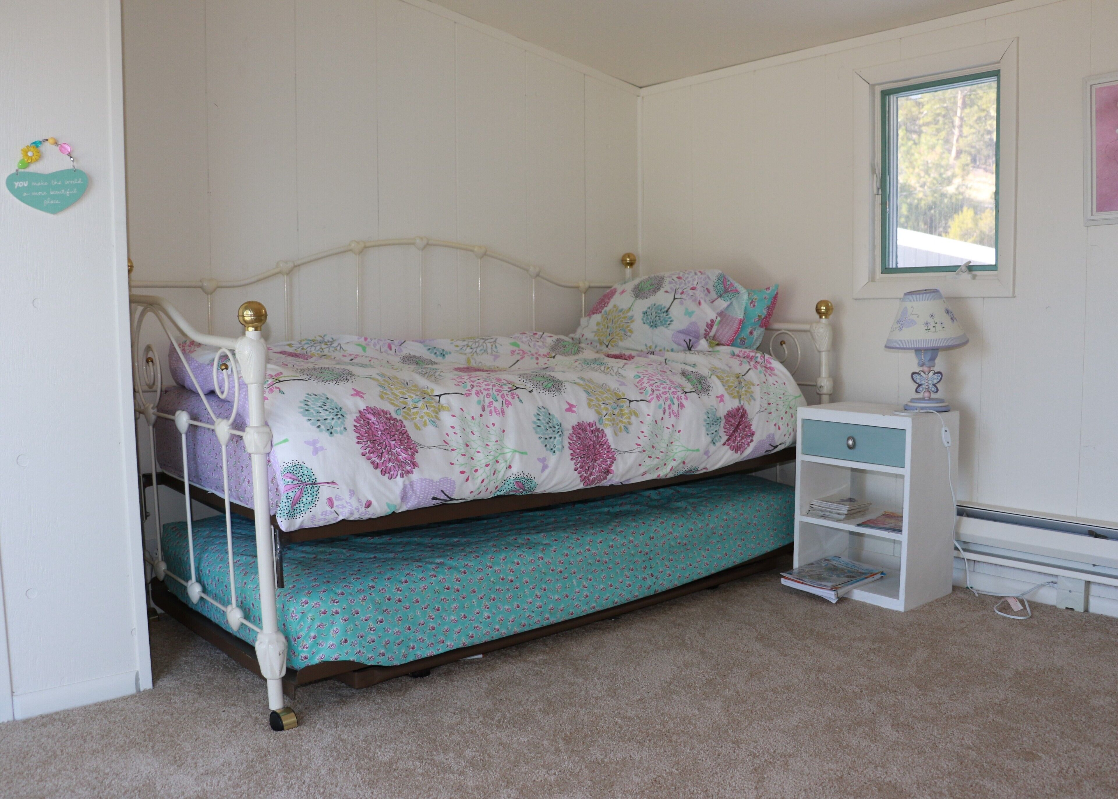 A single bed with a floral bedspread, a nightstand with a lamp, and a window with a view of trees.