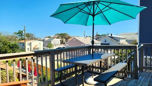 A wooden deck with a table and bench, a turquoise umbrella, and a view of houses in the background. - OCEANSIDE Getaway: Affordable 3 Bedroom, Top Floor Apartment with Pickleball Fun (Long Beach Township)