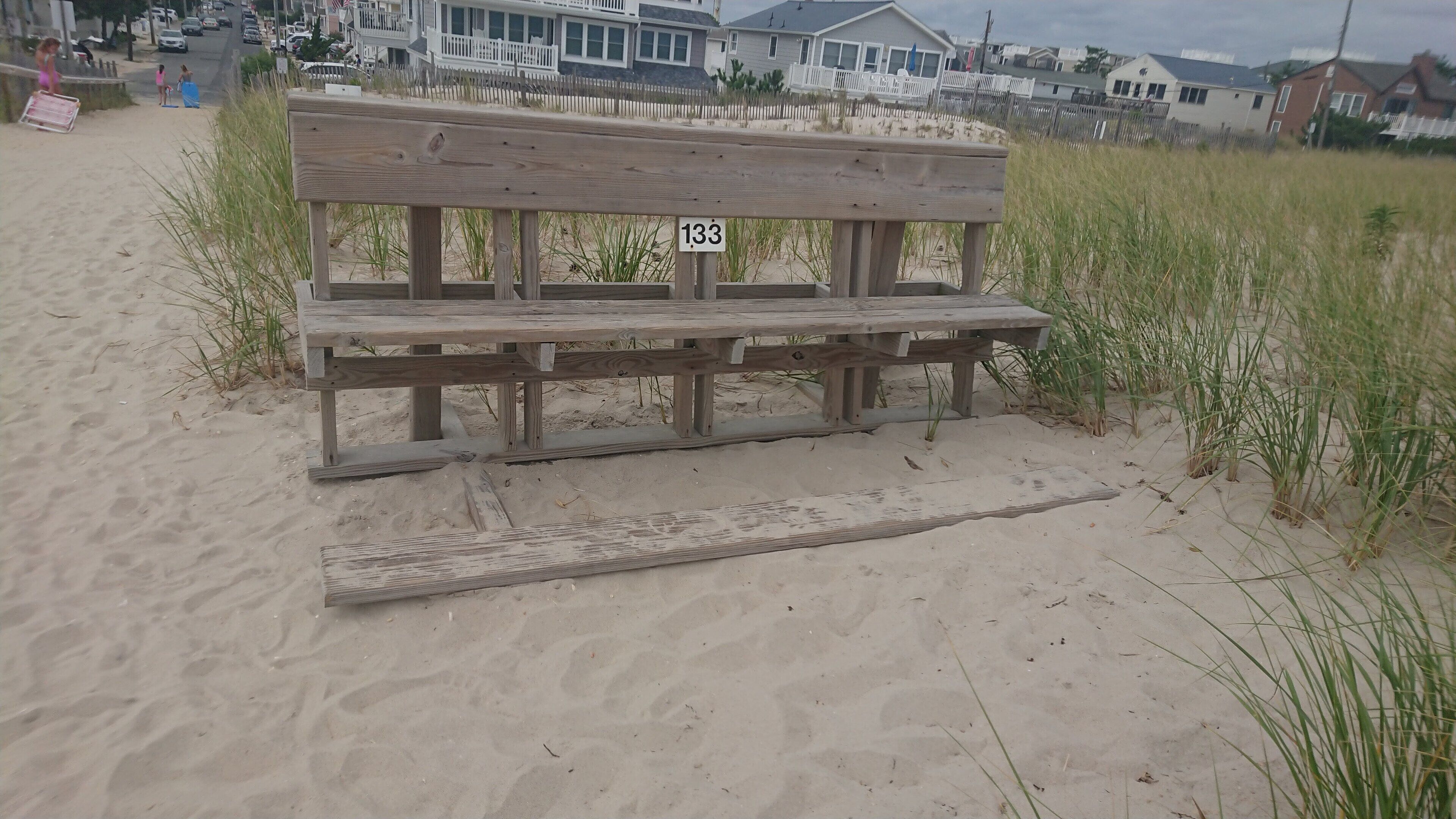 Una panca di legno con un cartello '133' su una spiaggia sabbiosa con erba alta.