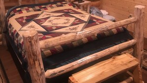 Un dormitorio de cabaña de madera con un marco de cama de madera, un edredón, una lámpara de noche y un cuadro en la pared.